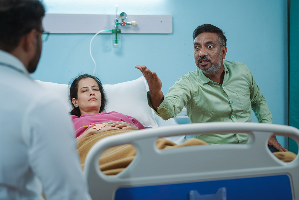 angry man next to a woman in a hospital bed arguing with a doctor
